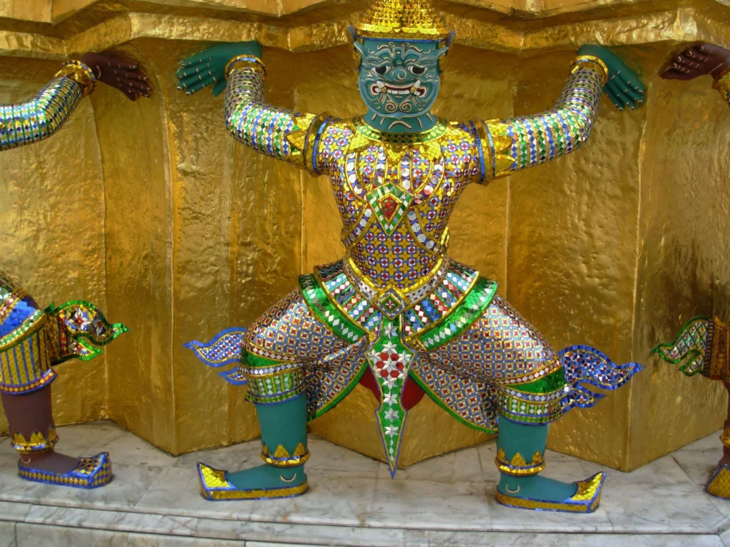 Colorful mosaic-covered Thai guardian statue with raised arms against a golden temple wall in Thailand.