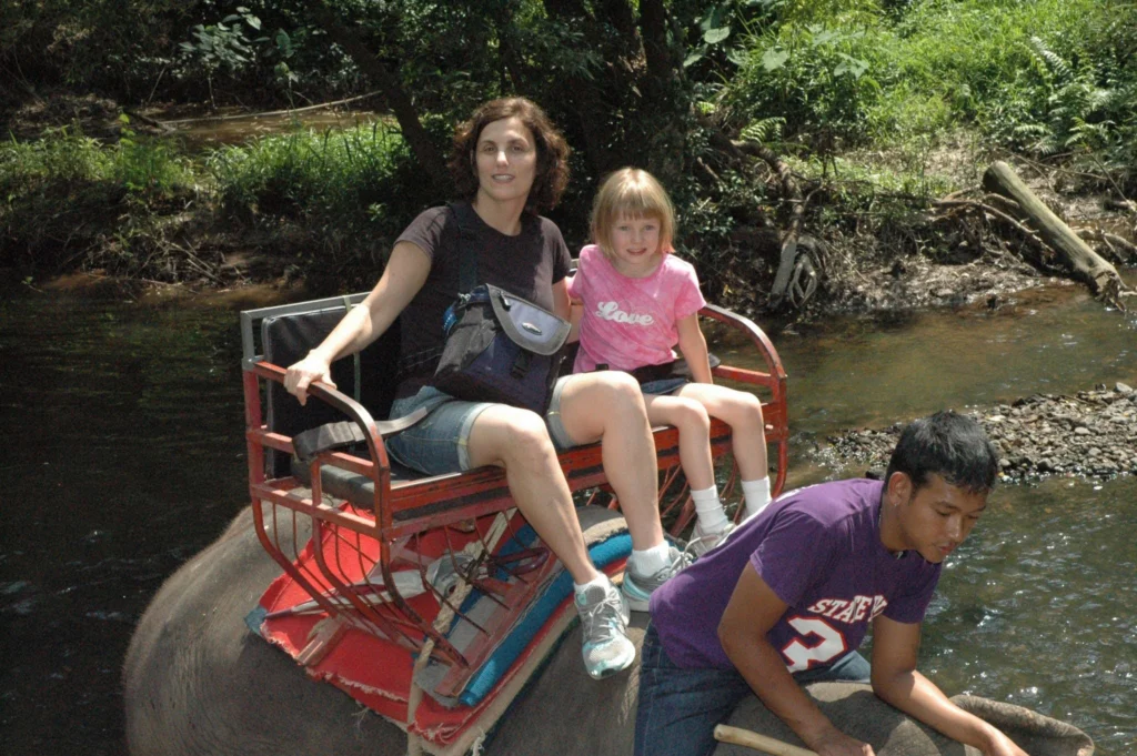Jessie as a child riding an elephant with an adult woman through a shallow river in Thailand.