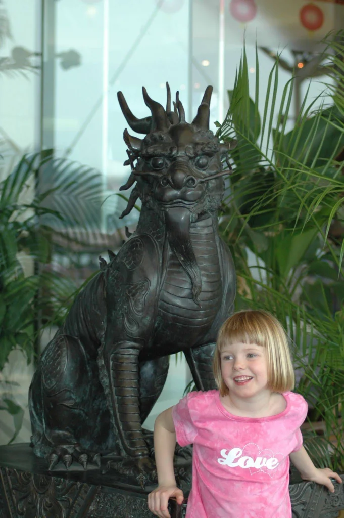 Jessie as a child standing beside a bronze dragon sculpture surrounded by tropical plants in Thailand.
